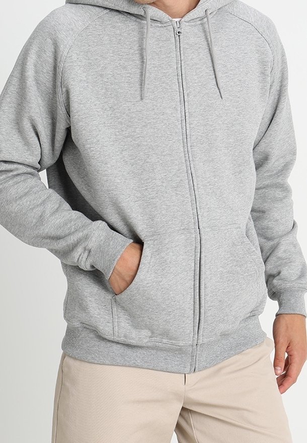 Personne portant un sweat à capuche gris clair zippé avec les mains dans les poches, associé à un pantalon beige, visible du milieu de la poitrine au milieu des cuisses.