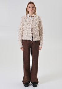 Cardigan crème à texture douce et finition duveteuse, associé à un pantalon marron à jambes larges et des chaussures plateformes noires. Design simple avec fermeture à boutons.