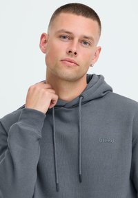 Joven con el cabello corto, vistiendo una sudadera gris con el logo "blend", sosteniendo el cuello cerca de su mentón, fondo neutro.