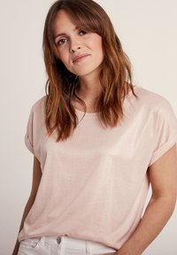 Helles rosa T-Shirt mit kurzen Ärmeln aus einem weichen, strukturierten Stoff. Rundhalsausschnitt und aufgerollte Ärmel. Tragen mit weißen Hosen.