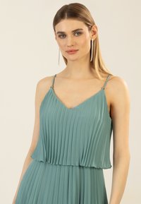 Faltkleid in hellblau mit V-Ausschnitt und dünnen Trägern, mit metallischen Akzenten an den Schultern. Glatter, fließender Stoff.