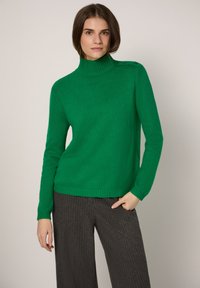 Grüner Strickrollkragenpullover mit geripptem Kragen und Bündchen. Verfügt über Knopfakzente an einer Schulter. Kombiniert mit dunklen, gestreiften, weit geschnittenen Hosen.