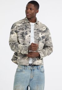 Camo-print jas in grijstinten en beige, met twee borstzakken, knoopsluiting en geborduurde branding op de voorkant. Draagt over een wit shirt.