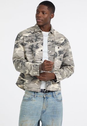 Camo-print jas in grijstinten en beige, met twee borstzakken, knoopsluiting en geborduurde branding op de voorkant. Draagt over een wit shirt.