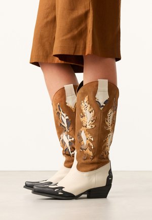 Legs wearing brown and cream cowboy boots with snake-print leaf designs and black pointed toes, paired with a rust-colored skirt.