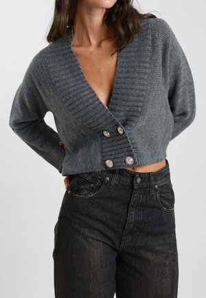 Cardigan grigio corto realizzato in tessuto a coste con scollatura a V profonda e due grandi bottoni metallici sul davanti. Indossato sopra jeans scuri.