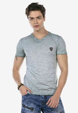 Graues V-Ausschnitt-T-Shirt mit einem strukturierten, verblassten Design und einem kleinen schwarzen Logo. Getragen mit blauen Jeans, die über Reißverschlussdetails verfügen.