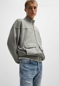 Grå zip-up jacka med två framfickor, utrustad med hög krage och ribbad nederkant, kombinerad med slitna blå denimjeans.