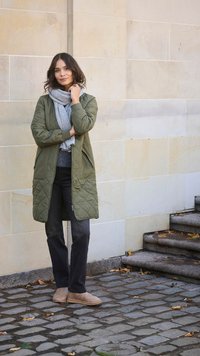 Quilted olive green long coat with two pockets, worn over a gray sweater and black jeans. Tan ankle boots and gray scarf complete the outfit.
