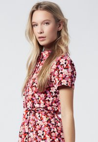 Abito floreale con uno sfondo rosso vivo decorato da fiori rosa e bianchi, maniche corte, colletto e una vita cintrata con una cintura per dare forma.