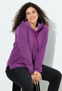 Donna sorridente con capelli ricci che indossa un felpa viola e jeans neri, seduta su uno sgabello nero contro uno sfondo bianco.