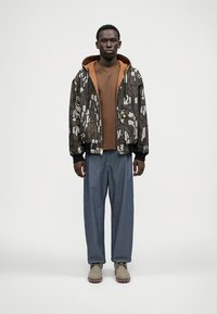 Braun-grau gemusterte Bomberjacke mit Kapuze über einem braunen Hemd, kombiniert mit blauen, weit geschnittenen Hosen und beigen Schuhen.