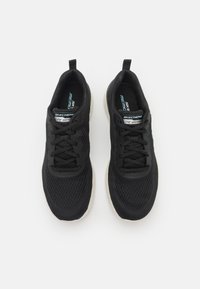 Sorte idrettssko med mesh-materiale, teksturert overdel og lysfarget såle. Funksjoner inkluderer snøring og merkevare på tungen.