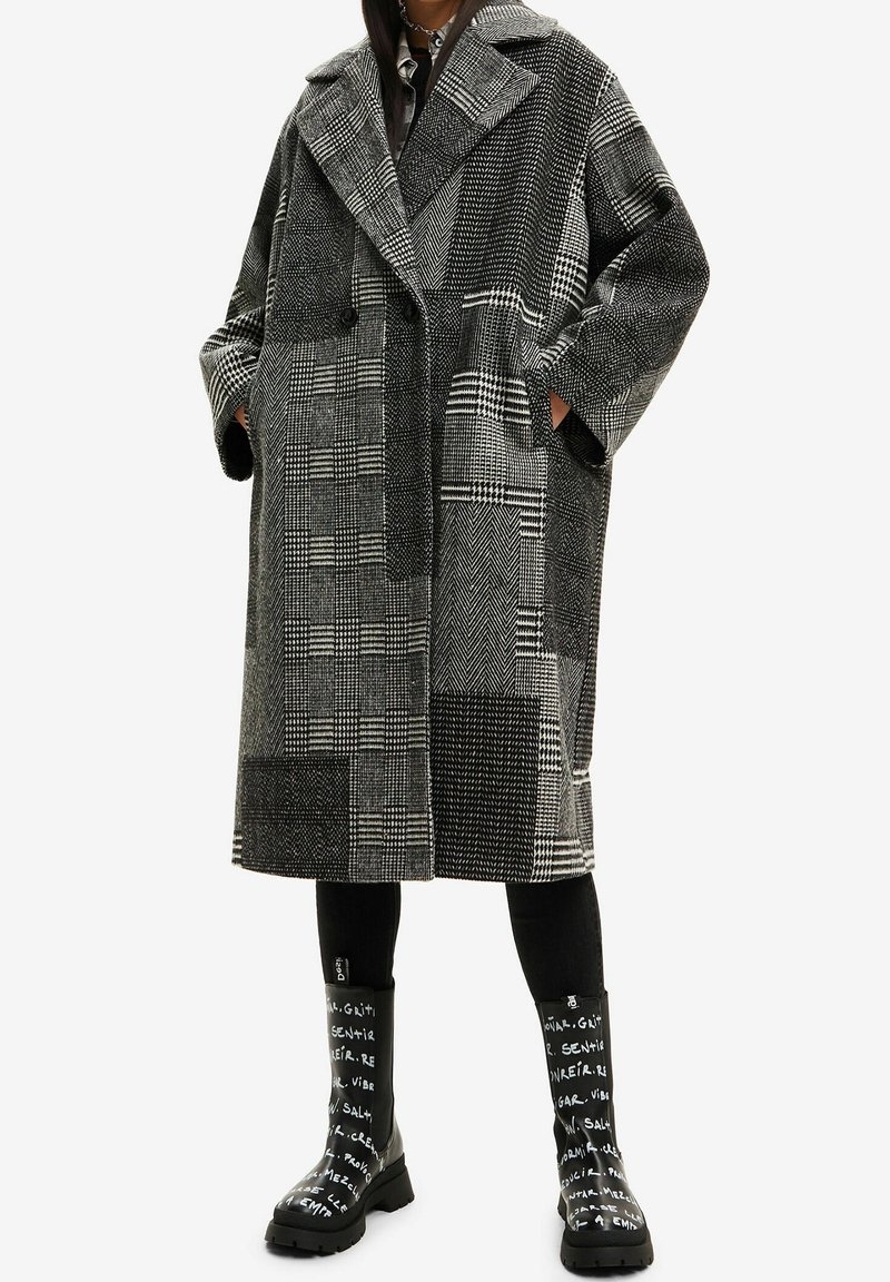 Femme portant un long manteau ample à motif noir et blanc, les mains dans les poches, leggings noirs et bottes noires avec texte blanc.