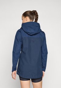 Marinblå vattentät jacka med en figursydd huva, långa ärmar och en något längre rygg. Har en diskret logotyp på övre ryggen.