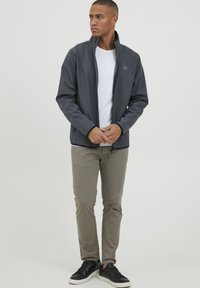 Gray, zip-up jacket with high collar, soft texture, paired with white t-shirt, light khaki pants, and black sneakers. Simple logo detail.