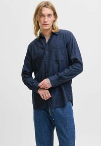 Blond man with tattoos wears a dark blue button-up shirt and blue jeans, standing with hands clasped against a plain background.