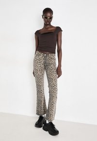 Bruin aansluitend shirt met korte mouwen, gecombineerd met leopardenprint flared pants. Zwarte chunky sneakers en olijfgroene zonnebril maken de look compleet.
