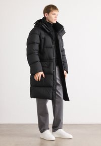 Sort polstret lang frakke med pelsforet krave, der har knaplukninger og lommer. Stylet over en sort turtleneck og grå bukser.