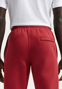 Pantalon de survêtement rouge avec une taille élastique, comprenant des poches latérales et une petite poche arrière. Fabriqué en tissu doux et épais.