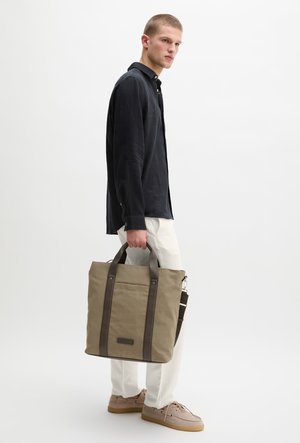 Jeune homme portant une chemise noire et un pantalon blanc, tenant un sac fourre-tout beige avec des anses foncées, debout devant un fond blanc uni.