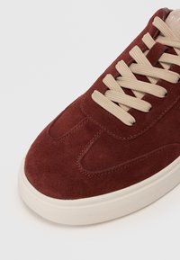 Primo piano di una sneaker in suede marrone con lacci beige e suola in gomma bianca e testurizzata su uno sfondo neutro.