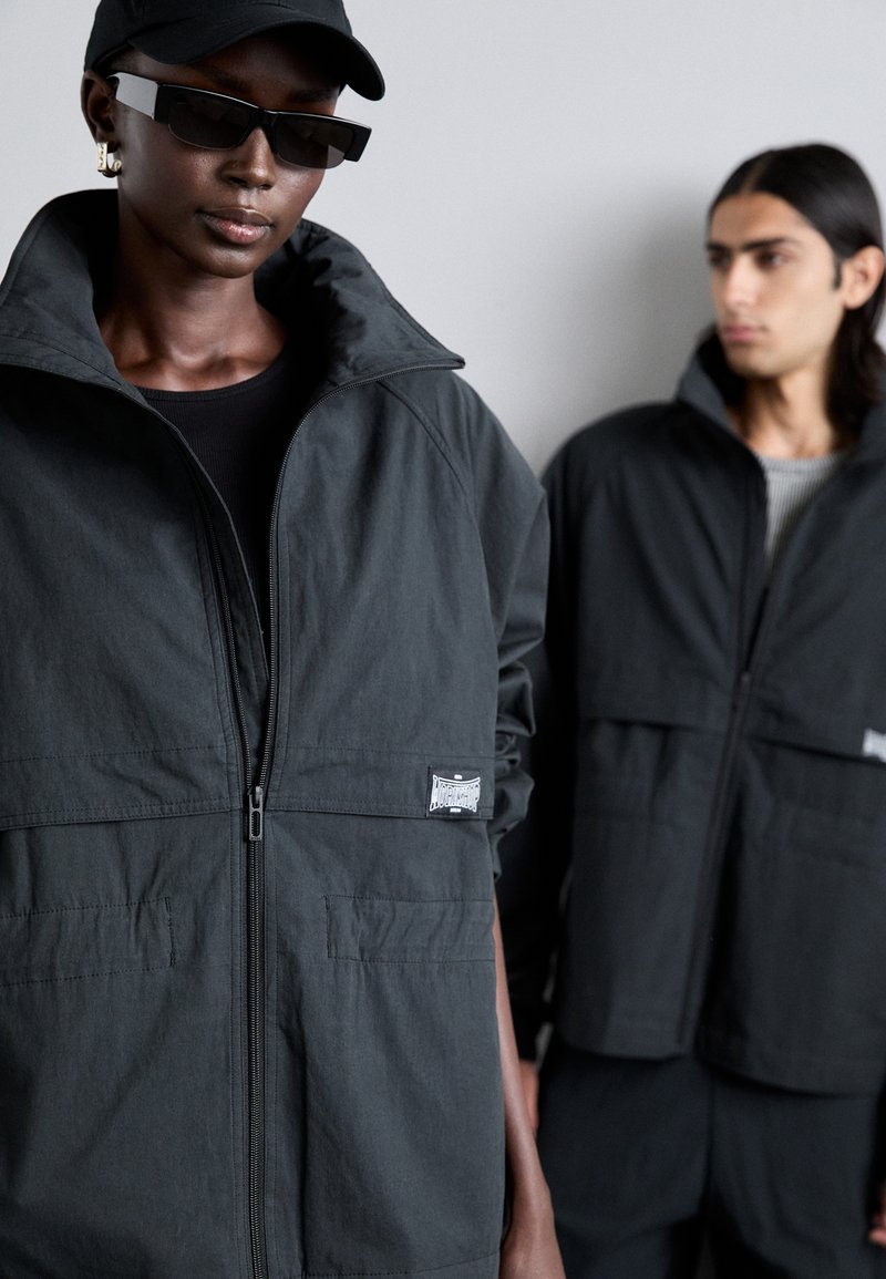 Chaqueta negra con cremallera y cuello alto, de tejido texturizado, con un detalle de parche en la parte inferior izquierda; combinada con una chaqueta de estilo similar al fondo.