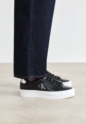 Person trägt schwarze Low-Top-Sneaker mit weißen Sohlen und "cK"-Logo, kombiniert mit dunkelblauen Straight-Leg-Denim-Hosen.