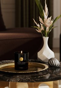 Czarna świeca z złotym emblematem oznaczonym "VELVET OUDH" na dekoracyjnym złotym tacy, otoczona białym wazonem i szarym kamieniem.