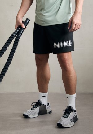 Uomo che indossa pantaloncini neri Nike, calzini bianchi Nike, scarpe da ginnastica grigie Nike, che tiene corde da battaglia nere in uno spazio interno neutro.