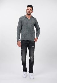 Homme portant un pull gris à torsades, un jean noir usé et des baskets blanches, debout devant un fond blanc uni.