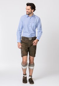 Homme portant une chemise à manches longues à carreaux bleu clair et un short en cuir brun avec des broderies décoratives, associé à des chaussettes à motifs arrivant aux genoux et des chaussures.