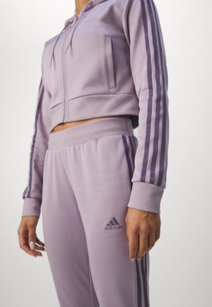 Personne portant un survêtement Adidas violet clair avec un sweat à capuche court zippé et un pantalon assorti avec trois bandes violet foncé et un logo.