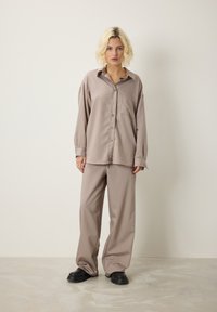 Giovane donna con capelli biondi corti, che indossa una camicia beige a righe sottili e pantaloni, abbinati a scarpe chunky nere, in piedi contro un muro chiaro e semplice.
