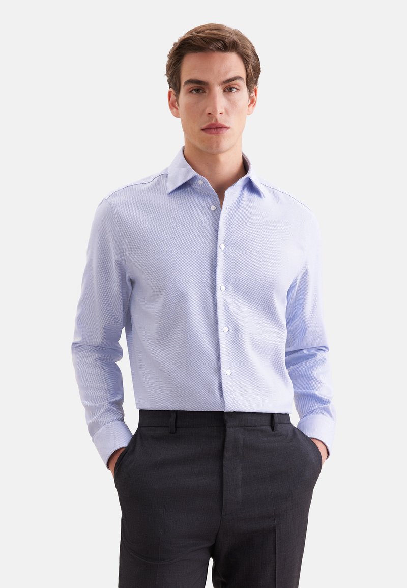 Homme portant une chemise bleu clair texturée et un pantalon gris foncé, les mains dans les poches, debout devant un fond blanc.