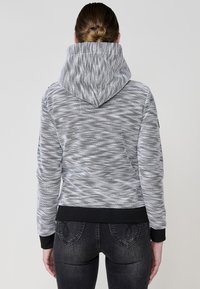 Szürke és fehér mintás kapucnis pulóver, tágas kapucnival, bordás fekete mandzsettákkal és derékrésszel, sötét farmernadrággal párosítva.