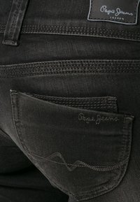 Vaqueros de denim negro con un bolsillo trasero decorado con costuras y un parche con el logo. La textura parece suave con un ligero brillo.