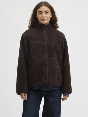 Femme portant une veste en polaire texturée brun foncé boutonnée, à manches longues et col haut, associée à un jean bleu foncé.