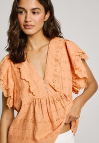 Blusa in cotone pesca con motivi a occhio, scollatura a V profonda, maniche ondulate e bottoni decorativi. Finitura testurizzata e vestibilità ampia.