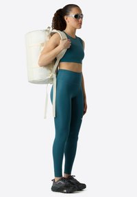 Un crop top de sport teal et un legging taille haute, portés avec un sac à dos cylindrique beige, comportant des bretelles ajustables et des chaussures noires.