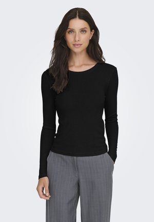 Mujer con cabello largo y oscuro que lleva una camiseta negra ajustada de manga larga y pantalones grises de rayas, de pie con una mano en el bolsillo.