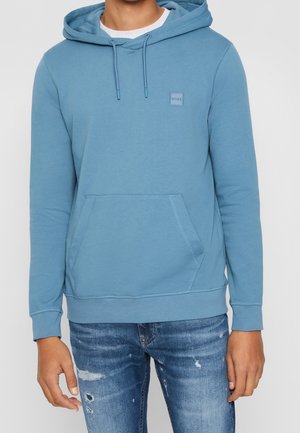 Uomo che indossa una felpa con cappuccio azzurro chiaro con tasca frontale e jeans blu sbiaditi con dettagli consumati.