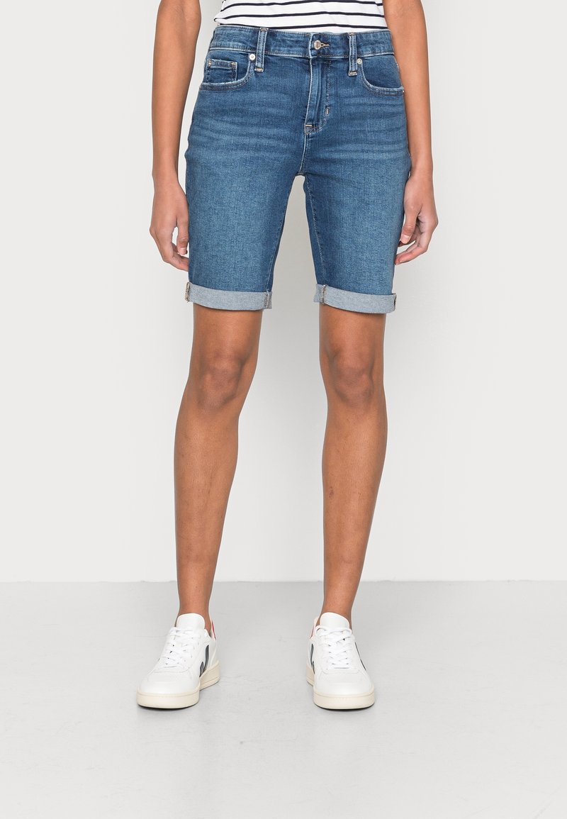 Personne portant un short en jean bleu retroussé à hauteur des genoux avec des poches avant et des baskets blanches, debout devant un fond clair uni.