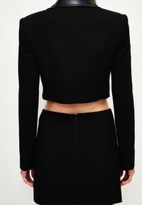 Blazer court noir à manches longues avec col en cuir, associé à une jupe noire taille haute dotée de poches et d'une fermeture éclair à l'arrière.