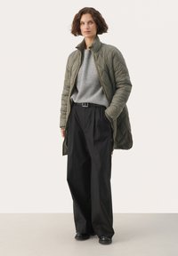 Vadderad olivgrön jacka med hög krage, grå rippad tröja och lösa svarta byxor. Svarta skor fulländar outfiten.