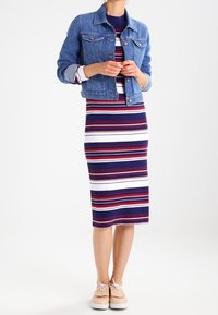 Giacca di jeans, abito midi a coste con strisce rosse, bianche e blu, e scarpe beige con dettagli a fiocco. Texture lisce e design aderente.