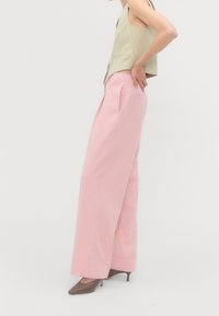 Femme portant un pantalon rose à jambes larges, un haut beige sans manches et des talons pointus marron, debout de profil avec une main sur la hanche.