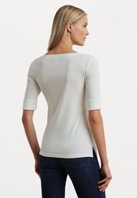 Λευκό, εφαρμοστό πουκάμισο με τρία τέταρτα μανίκια και διπλωμένα μανίκια. Λείος υφάνσεις, φαρδύς λαιμός και πλαινά ανοίγματα στο ύψος του στριφώματος.