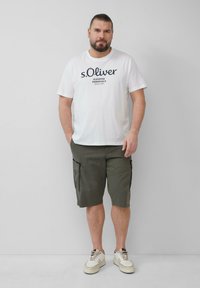 Biała bawełniana koszulka z granatowym napisem, zielone szorty cargo z bocznymi kieszeniami oraz beżowe trampki z subtelnymi akcentami i teksturowanymi podeszwami.