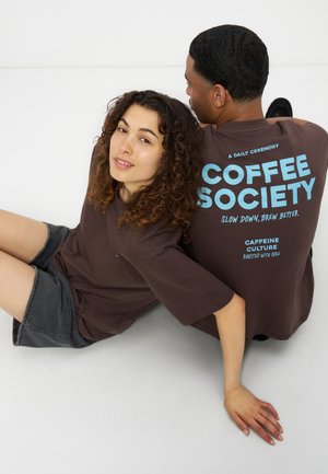 Deux personnes sont assises dos à dos sur le sol, portant des chemises marron avec un texte bleu « Coffee Society » et des shorts décontractés sur fond blanc.